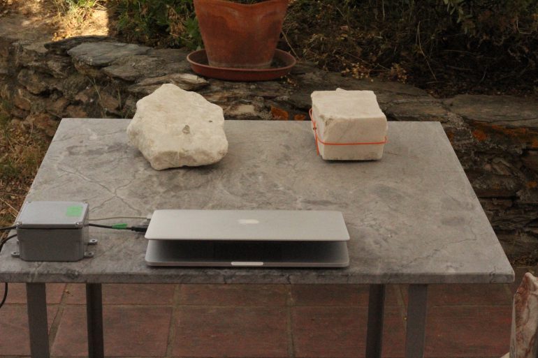 On an outdoor terasse a rectangular marble table are placed two blocks of marble, a computer and a grey box which is connected to the computer. One marble block has an orange plastic strip around it. The grey box has a green piece of tape. 