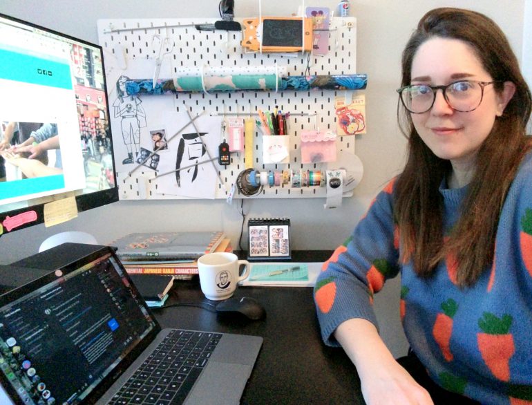 gina poses in front of a laptop desk and a wall panel full of craft supplies